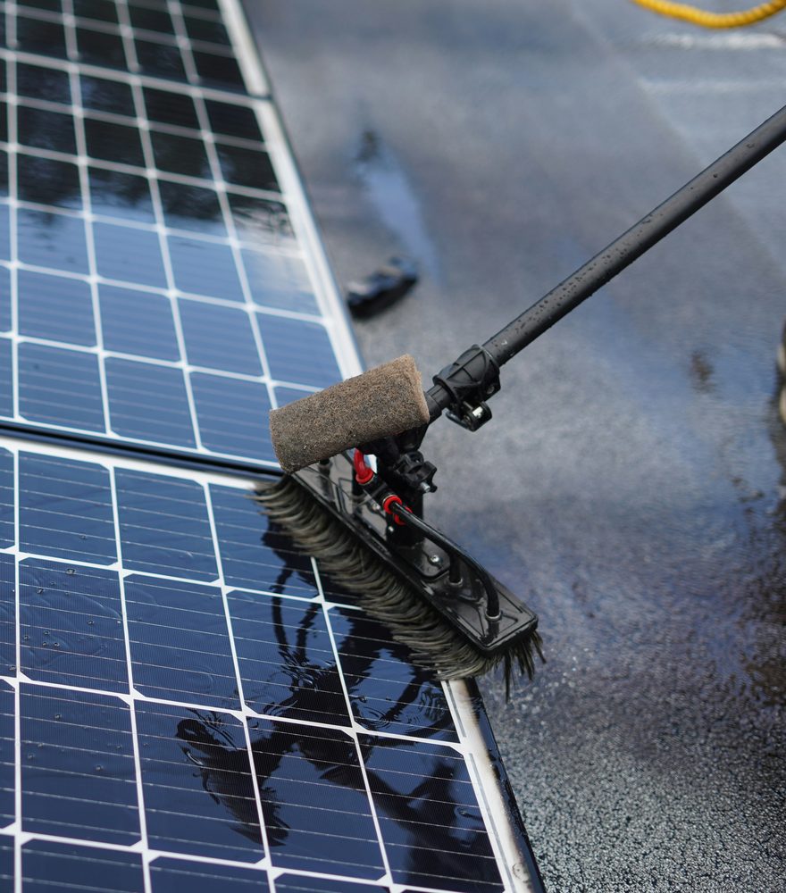 A technician cleaning a rooftop solar panel with a wet brush on a telescopic pole